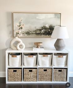 a white shelf with baskets on it and a painting in the corner behind it next to a lamp