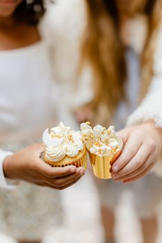 two women holding cupcakes with white frosting on them and gold foil wrappers