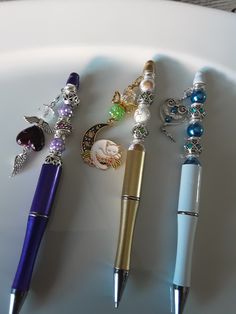 three pens are lined up next to each other on a white plate with beads and charms