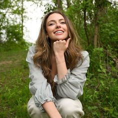 a woman sitting in the grass with her hand on her chin and smiling at the camera