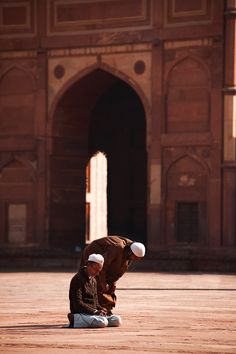 two men sitting on the ground in front of a building with arches and doorways