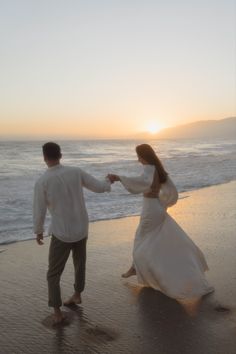 a man and woman walking on the beach at sunset holding hands with each other,