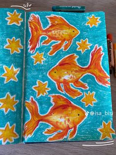 an open notebook with goldfish and stars on it, next to crayon markers