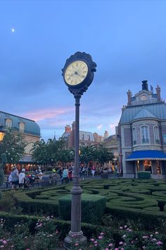 a large clock on top of a metal pole in the middle of a garden area