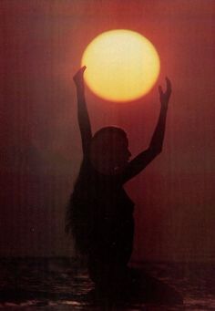 a woman is standing in the water with her arms up as the sun goes down