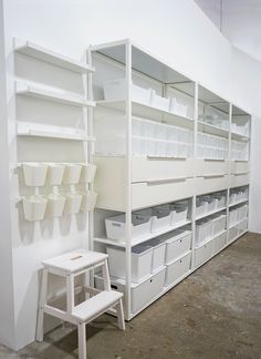 an empty room with white shelves and plastic bins on the wall next to a step stool