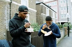 two young men eating hot dogs on the sidewalk in front of a brick building, one is wearing a black hoodie and the other wears a blue sweatshirt