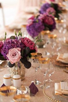 the table is set with purple and pink flowers in vases, wine glasses, and napkins