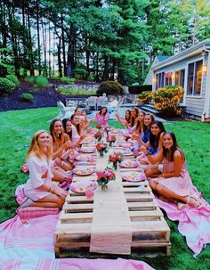 a group of women sitting around a wooden table eating food on pink blankets in front of a house