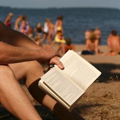 a man reading a book on the beach while others are in the water behind him