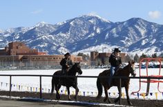 two men riding horses in the snow near a fence with mountains in the back ground