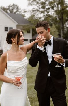 a bride and groom standing in front of a white house holding wine glasses with their hands