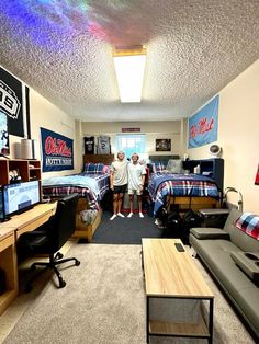 two men are standing in the middle of a dorm room with beds and desks