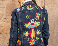 a woman wearing a denim jacket with colorful embroidered designs on the back and sides, standing in front of a stone wall