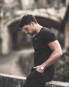 a man in black shirt and sunglasses leaning on wall with his hand on his hip