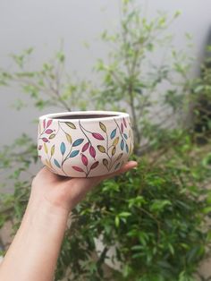 a hand holding up a ceramic planter in front of a tree with leaves on it