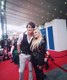 a man and woman standing on a red carpet in front of a stage with people