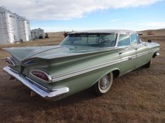 an old green car parked in the middle of a dry grass field next to a barn