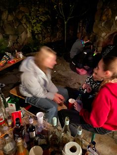 two people sitting at a table with bottles and cups on it, one person is talking to the other