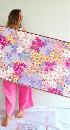 a woman in pink pants holding up a large floral painting on a white wall above her head