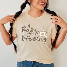 Celebrate the sweetest news with our adorable Pregnancy Announcement Shirt! This charming tee features the phrase A Baby is Brewing elegantly written across the front, capturing the excitement of a new addition to your family. A whimsical pumpkin, adorned with delicate baby footprints, adds a heartwarming touch to the design. Crafted with soft, high-quality fabric, this shirt ensures comfort and style as you share the joyous journey ahead. Whether you're announcing to friends, family, or the world, this shirt is a delightful way to spread the news of your growing pumpkin patch! Bella + Canvas t-shirts and Gilden 18000 heavyweight sweatshirts are featured in my store. They are buttery soft, smooth and so comfy. They are a tried and true favorite of mine, and are sure to be yours as well.  B Cute Maternity T-shirt With Graphic Print, Cute Maternity Graphic Print T-shirt, Cute Maternity T-shirt With Letter Print, Cute Maternity Cotton T-shirt, Maternity Tees, Pregnancy Announcement Shirt, Pregnancy Humor, Xmas Shirts, Dog Lover Shirt