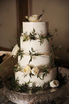 a three tiered cake with white flowers and greenery on the top is sitting on a silver platter