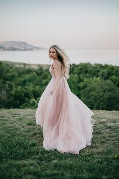 a woman in a pink dress standing on top of a lush green field next to the ocean