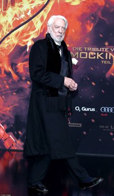 an older man in a long black coat and tie walking on a red carpet with flames behind him