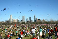 a large group of people flying kites in a park with tall buildings behind them