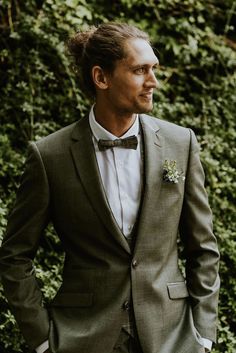 a man wearing a suit and bow tie standing in front of some bushes with his hands in his pockets