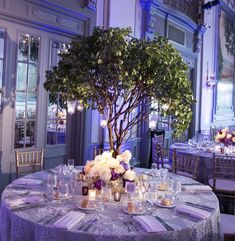 the table is set for an event with white flowers and candles on it in front of a tree