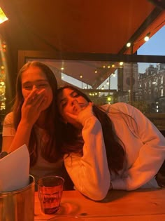 two young women sitting at a table with drinks in front of them, both covering their mouths