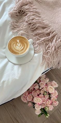 a cup of coffee sitting on top of a bed next to pink flowers and a blanket