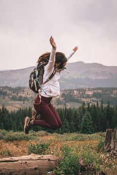 a woman jumping in the air with her backpack over her head and mountains in the background