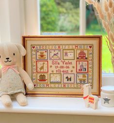 a teddy bear sitting next to a framed cross - stitch pattern in front of a window