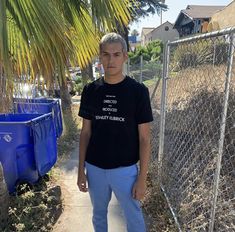 a man standing on a sidewalk next to a chain link fence and blue bins