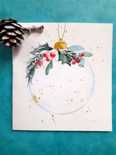 a watercolor christmas ornament with holly and pineconis on white paper