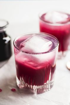 two glasses filled with red liquid on top of a white tablecloth next to silver spoons