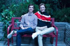 two people sitting on a red bench in front of some bushes and trees, one person is smiling at the camera