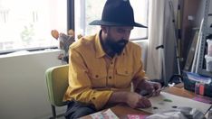 a man sitting at a desk in front of a window wearing a black hat and yellow shirt