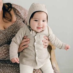 A baby, dressed in a Henry linen shirt and white pants made from a linen/cotton blend, is being held by a woman wearing a floral dress. The happy child sits on a wooden chair set against a light background. Beige Long Sleeve Tops For Playtime, Beige Cotton Tops For Playtime, Cream Tops For Spring Playtime, Flax Cotton Button-up Tops, Flax Color Cotton Button-up Tops, Flax-colored Cotton Button-up Tops, Cream Linen Loungewear Tops, Cream Linen Tops For Loungewear, Long Sleeve Shirt For Spring Playtime