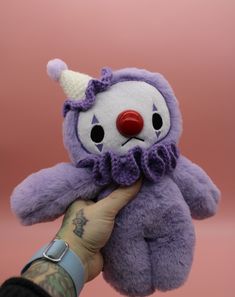 a hand holding a purple stuffed animal with a clown face on it's head