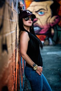 a woman leaning against a brick wall wearing sunglasses and a hat