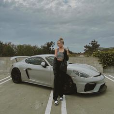 a woman standing next to a silver sports car in a parking lot with her hand on the door handle