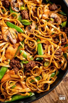 a skillet filled with noodles, meat and veggies on top of a wooden table