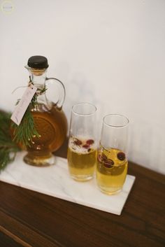 two glasses and a bottle of alcohol on a wooden table next to a pine branch