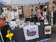 people are standing under a tent at an outdoor flea market with shirts and hats on display