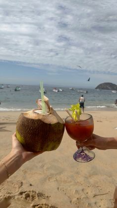 two people toasting on the beach with cocktails in their hands and one holding up a coconut