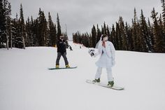 two snowboarders are standing in the snow near some trees and one is wearing a white coat