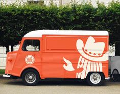 an orange and white food truck parked in front of a building next to a hedge